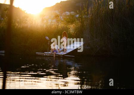I bambini possono godersi una tranquilla giornata di pesca su un molo di legno, inondato dalla luce dorata del tramonto. L'acqua serena riflette la loro gioia e il rigoglioso re circostante Foto Stock