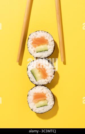 Vista dall'alto di tre deliziosi sushi roll con salmone e cetriolo, elegantemente posizionati con bacchette di legno appuntite su un vivace sfondo giallo, Foto Stock