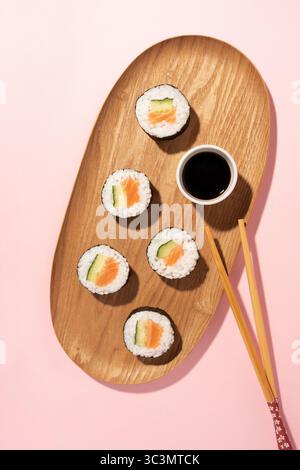 Vista dall'alto dei sushi roll con cetriolo e salmone su un piatto di legno, accompagnati da salsa di soia e bacchette, su sfondo rosa. Un giapponese Foto Stock