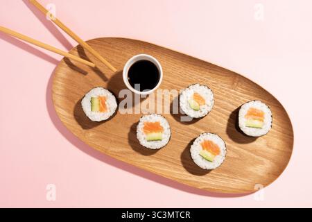 Vista dall'alto dei sushi con salmone e cetriolo su un piatto di legno, accompagnati da bacchette e salsa di soia, su uno sfondo rosa pastello in una s. Foto Stock