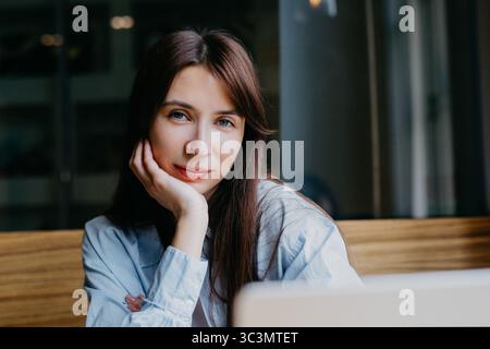 Donna seduta in un bar, che lavora al suo notebook con un'espressione premurosa. E' vestita con un abbigliamento casual, la sua mano poggia sul mento, circondata Foto Stock
