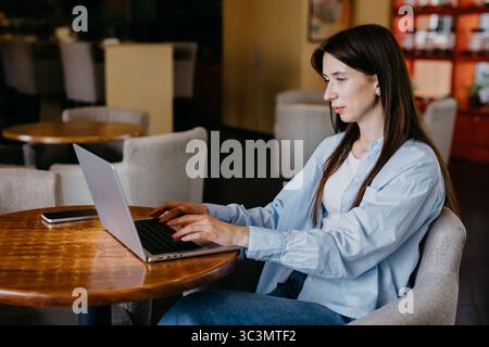 Una donna lavora sul suo computer portatile in un accogliente bar, circondato da comode sedie e da un'illuminazione soffusa. La sua attenzione e la sua produttività sono evidenti nell'invito Foto Stock
