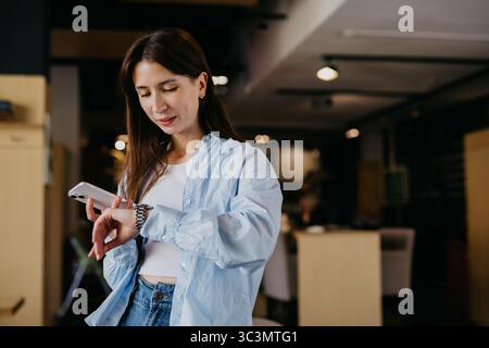 Una donna che indossa una camicia blu controlla il suo orologio mentre tiene in mano uno smartphone in un ambiente accogliente. Sembra concentrata, gestendo il suo tempo e il suo lavoro in modo scheggiato Foto Stock