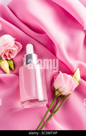 Un'elegante bottiglia cosmetica con contagocce poggia su morbide pieghe di seta rosa, accompagnate da eleganti fiori di eustoma, creando un atmos lussuoso e sereno Foto Stock