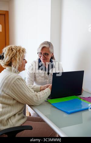 Un'immagine gioiosa di due amiche anziane sedute comodamente a casa, che si impegnano in una conversazione vivace mentre si utilizza un notebook sul tavolo, sorridendo Foto Stock