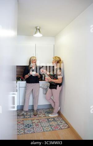 Due amiche anziane condividono un momento di gioia con un caffè in una cucina moderna. Stanno chiacchierando e sorridendo, si godono la compagnia reciproca e il Foto Stock