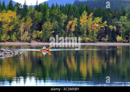 Un kayak solitario scivola attraverso la superficie vetrata del lago McDonald, incorniciata da riflessi della foresta autunnale e da lontane vette montane sotto un cielo ripido. Foto Stock