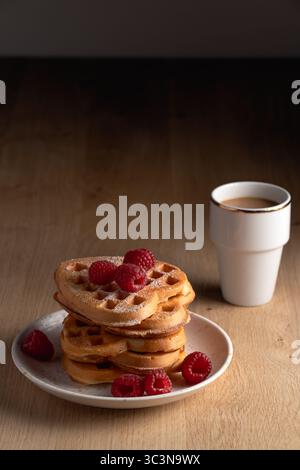 Un ambiente incantevole con una pila di waffle a forma di cuore, lamponi freschi e una tazza di caffè bianco, perfetto per una colazione accogliente Foto Stock