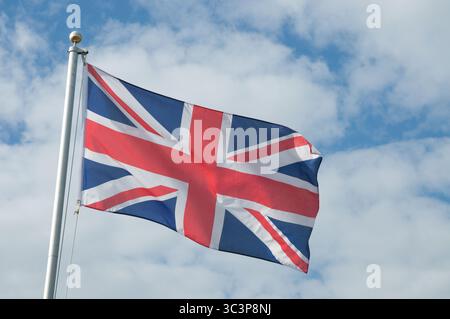 La bandiera Union Jack sventola orgogliosamente su un palo, posto su uno sfondo di nuvole sparse e luce solare brillante durante il giorno. Foto Stock