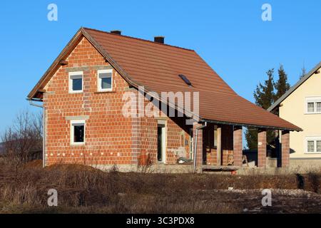 Una casa in mattoni parzialmente costruita con un tetto in piastrelle rosse inclinate sorge in un'area rurale erbosa, caratterizzata da muratura a vista e pareti non dipinte sotto Foto Stock