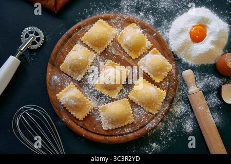 Una splendida disposizione di ravioli appena fatti su una tavola di legno circondata da farina, uova e utensili da cucina pronti per una deliziosa creazione di pasta Foto Stock
