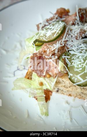 Panino aperto a strati con ingredienti freschi, carni salate, verdure croccanti e condimento con formaggio grattugiato per un'esplosione saporita Foto Stock