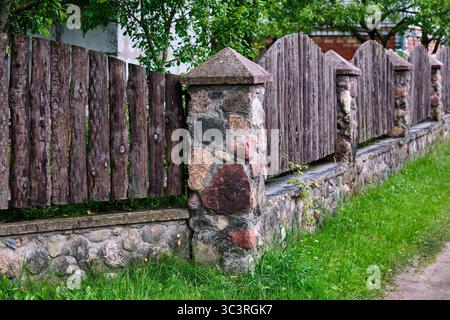 Una caratteristica recinzione in legno fatta di tavole intemperie si erge accanto ad un muro di pietra, circondata da lussureggiante vegetazione in una tranquilla area di campagna. Foto Stock