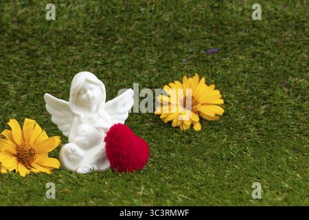 Una figura d'angelo con un cuore rosso e fiori gialli sull'erba verde Foto Stock