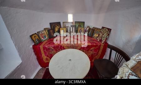Un tavolo con coperte rosse e diverse icone fiancheggiate da candele in una piccola stanza, cappella, chiesa, Agios Isidoros, isolotto roccioso, Gourna Bay, Kokkali, Lero Foto Stock