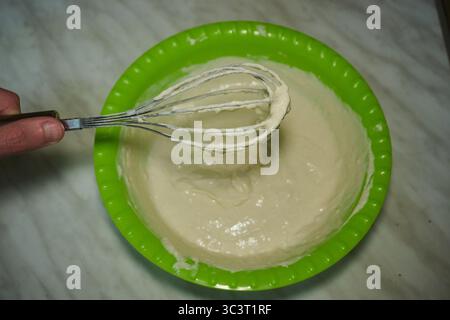 Frusta per montare l'impasto con una frusta in un recipiente verde sul ripiano della cucina. Foto Stock