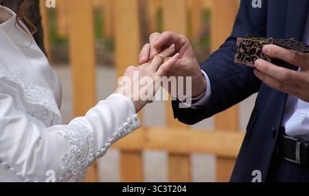 La giovane coppia al matrimonio scambia anelli di fidanzamento. Le coppie della coppia sono a contatto, gli anelli di innesto dorati brillano contro il backgrou Foto Stock