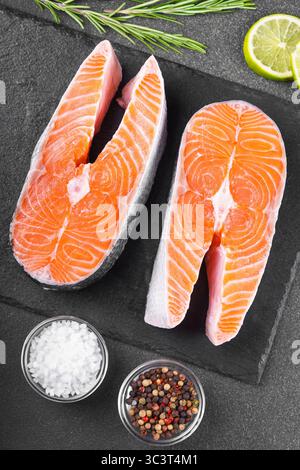 Due bistecche di salmone o trote fresche su una superficie di pietra nera con ingredienti alimentari. Pomodori, lime, rosmarino, sale e pepe. Mangiare sano, pesce Foto Stock