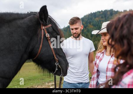 Il giovane uomo e la donna che indossano un cappello da cowboy guardano un cavallo scuro in piedi in un campo erboso. Gli alberi stanno sullo sfondo. Foto Stock