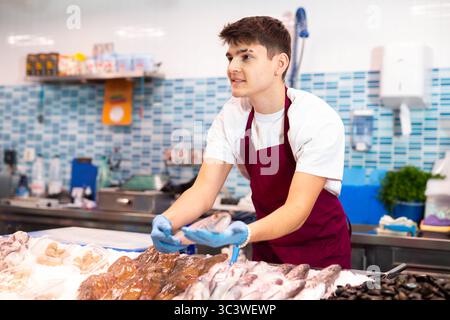 Venditore positivo in grembiule in piedi vicino al banco che offre pesce striato triglie rosse Foto Stock