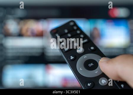 Una mano che tiene in mano il telecomando commuta i canali di un televisore con uno schermo sfocato sullo sfondo. Le riprese simboleggiano il moderno consumo dei media, il sovraccarico di contenuti, l'affaticamento delle scelte digitali e la costante ricerca di informazioni coinvolgenti. Ideale per temi riguardanti le abitudini multimediali, l'intrattenimento, l'età dell'informazione e la dipendenza dallo schermo. Foto Stock
