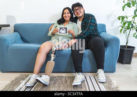Donna incinta con gamba protesica seduta sul divano con marito, che mostra un'immagine ecografica del bambino Foto Stock