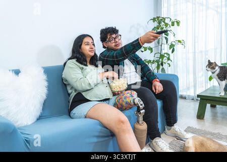 Giovane coppia amorevole che si rilassa sul divano, guarda la televisione e mangia popcorn, accompagnati dai loro animali domestici Foto Stock