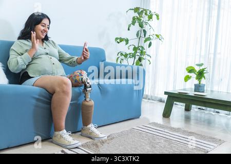 Donna incinta felice con protesi che ondeggia la gamba mentre effettua una videochiamata utilizzando lo smartphone in salotto Foto Stock