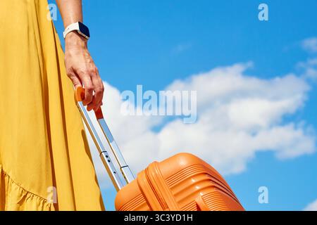 Donna in abito giallo che tiene in mano una valigia arancione contro il cielo blu con nuvole. Primo piano di una donna viaggiatrice con bagagli. Concetto di viaggio elegante, va Foto Stock