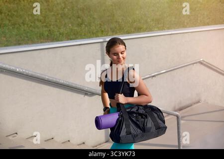 Giovane donna che porta borsa da palestra e tappetino yoga che scende le scale in un ambiente urbano stile di vita sano concetto di vita sano. Foto Stock