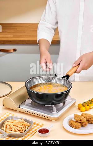 Preparazione di deliziosi piatti fritti con filtro a rete in acciaio inossidabile e piano cottura moderno, cuoco che cucina pepite di pollo in una padella su un gattino Foto Stock