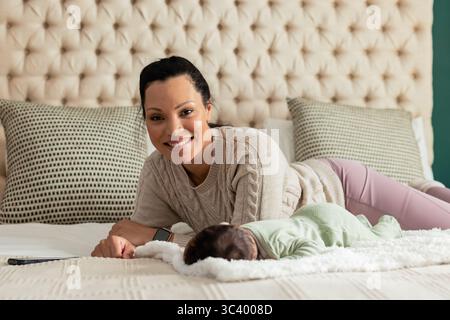 Madre afroamericana sdraiata sul letto di casa vicino al neonato in coperta, sorridente, che raggiunge lo smartphone Foto Stock