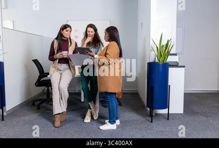 Diverse collaboratrici che tengono la tazza di caffè e puntano il tablet con appunti sulla lavagna in ufficio Foto Stock