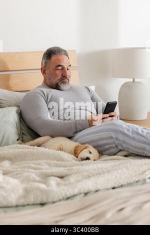 Uomo seduto sul letto con pigiami e smartphone a scorrimento accanto al cucciolo e alla lampada in ceramica Foto Stock