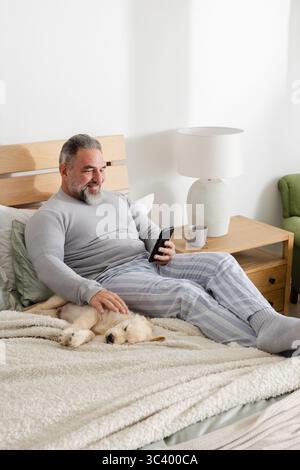 Uomo anziano seduto sul letto con pigiama che tiene in mano lo smartphone e accarezza il cucciolo sotto la coperta Foto Stock