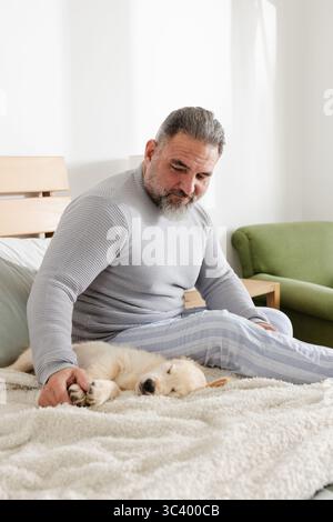 Uomo anziano che indossa un maglione grigio e pigiama a righe seduto sul letto in camera da letto che accarezza il cucciolo Foto Stock