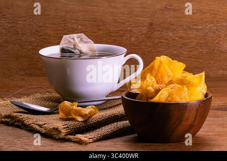Patatine dolci e croccanti in una ciotola di legno e una tazza di tè con bustina di tè in una tazza di ceramica bianca e un cucchiaio di metallo su un panno da sacco posto su un tavolo di legno. Foto Stock