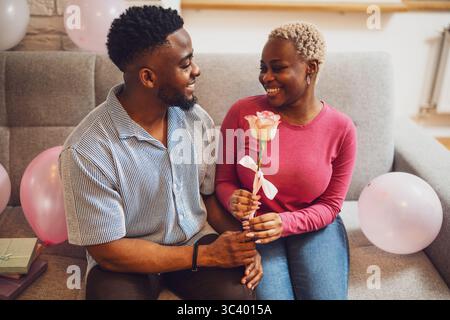Felice coppia amorevole che festeggia il compleanno della donna a casa. Il fidanzato sta dando la rosa alla sua ragazza. Foto Stock