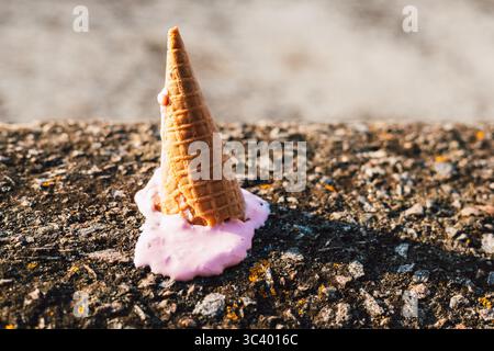 Cono gelato caduto con gelato rosa versato su una superficie di cemento testurizzato. Un cono gelato, rovesciato su una superficie ruvida di pietra con versamenti Foto Stock