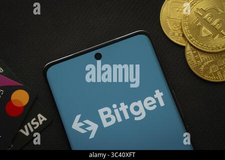 Dhaka, Bangladesh- 27 luglio 2025: Smartphone con logo Bitget circondato da monete e carte di pagamento Bitcoin Foto Stock