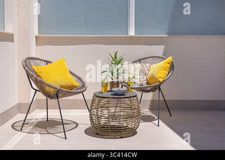 Patio elegante e accogliente con due sedie rotonde con cuscini gialli, un tavolo in vimini, bevande e un pezzo centrale della pianta. Foto Stock