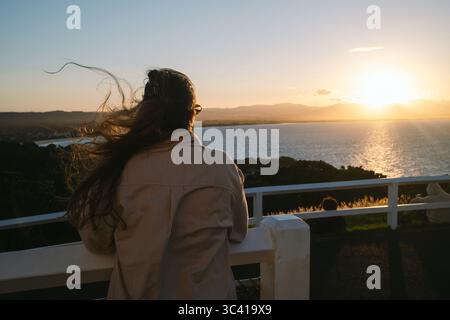 Donna guarda Byron Bay tramontare sull'oceano dalla ringhiera panoramica. Foto Stock