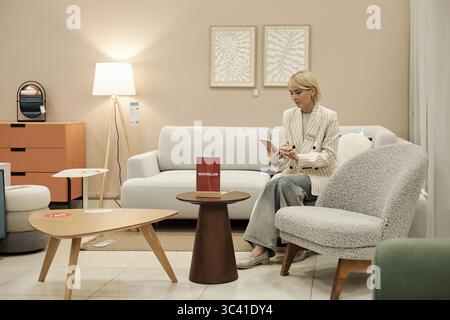 Donna caucasica seduta sulla sedia leggendo brochure nello showroom di un negozio di mobili circondata da tavoli e sedie moderni, incentrata sulle informazioni sui prodotti, sfondo neutro Foto Stock