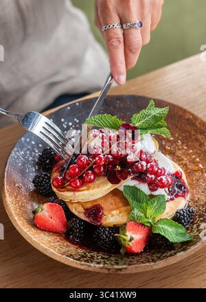 Primo piano di una persona che taglia una pila di pancake conditi con frutti di bosco freschi e panna utilizzando una forchetta e un coltello. Colazione accogliente o brunch. Foto Stock