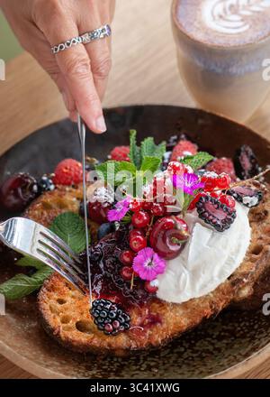 Primo piano di una persona che taglia una fetta di pane tostato ricoperto di frutti di bosco freschi e panna utilizzando una forchetta e un coltello. Colazione salutare o brunch fatto in casa. Foto Stock