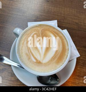 Vista dall'alto verso il basso di una tazza per cappuccino di colore chiaro, appoggiata su un piattino coordinato con superficie che presenta una latte art ben eseguita a forma di cuore. Foto Stock
