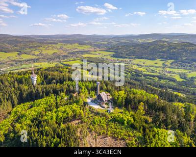 Una vista aerea cattura la torre di osservazione di Svatobor circondata da lussureggianti foreste verdi e colline ondulate in Cechia. Il paesaggio mostra la bellezza della natura sotto un cielo azzurro. Foto Stock