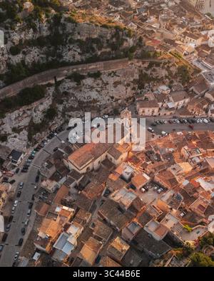Vista aerea dei tetti in terracotta che scendono fino alla chiesa barocca, incorniciata da aspre scogliere e strade tortuose, Scicli, Sicilia, Italia. Foto Stock