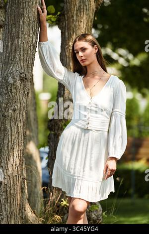 Una giovane donna posa elegantemente in un affascinante vestito bianco vicino a un albero, crogiolandosi alla luce calda del sole. Foto Stock
