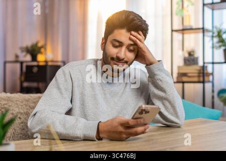Giovane indiano seduto sul divano a tavola, scrivendo attivamente sullo smartphone, appoggiando la testa a mano nel soggiorno di casa. Il ragazzo arabo sembra concentrato, profondamente impegnato nella conversazione o nel compito Foto Stock